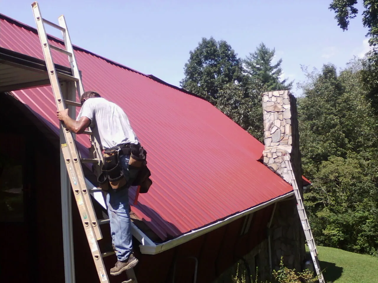 Roofer on a ladder beside a red metal roof and stone chimney — Soffit & Fascia Repair by Roofing Brothers in East Greenville